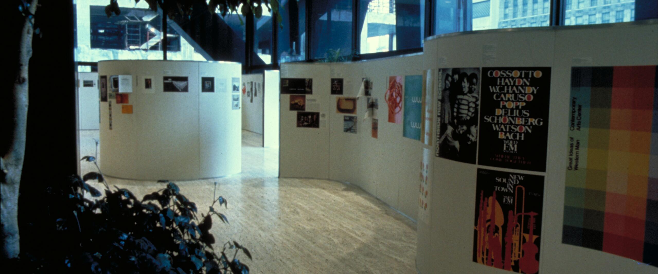 Curved indoor gallery space with posters displayed on white walls. Large windows in the background show a view of buildings outside. A leafy plant is visible in the foreground.