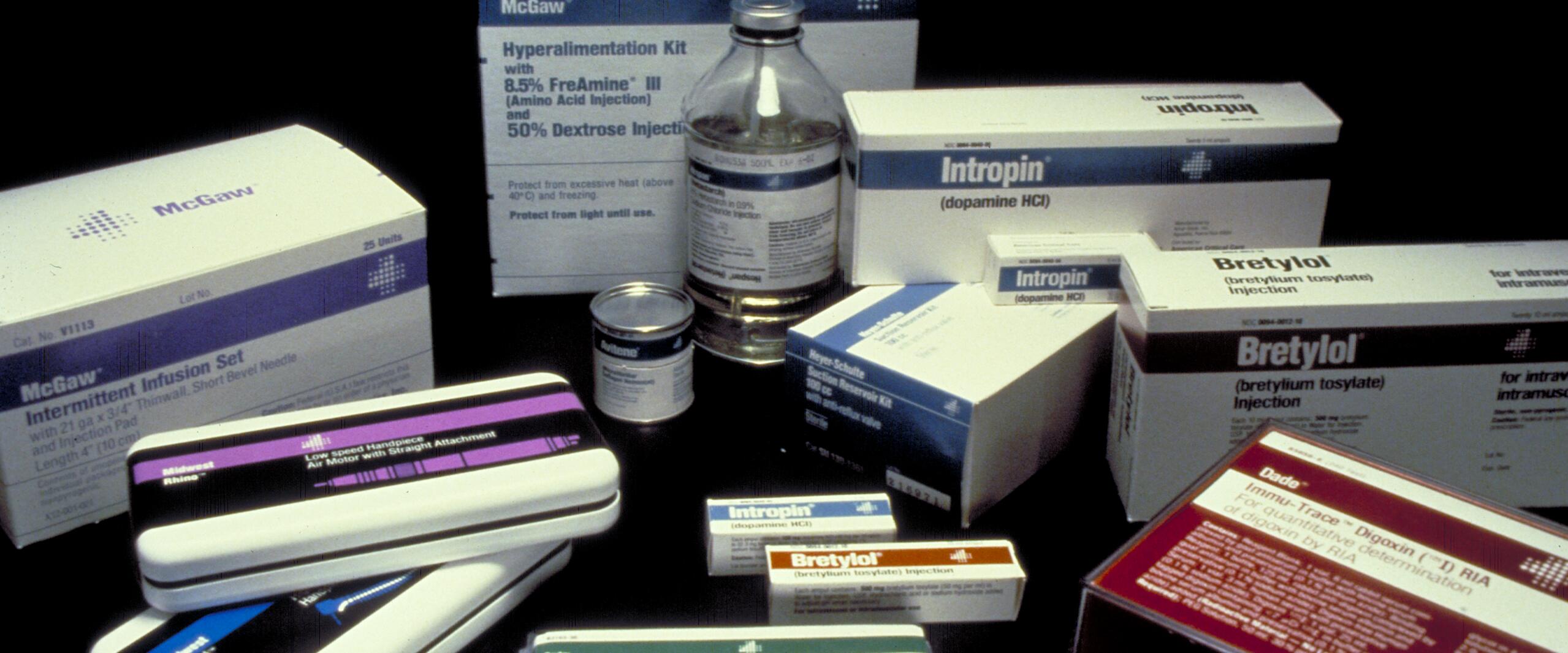 Various medical supply boxes and vials arranged on a dark surface, including packages labeled Bretylol, Intropin, and a hyperalimentation kit.