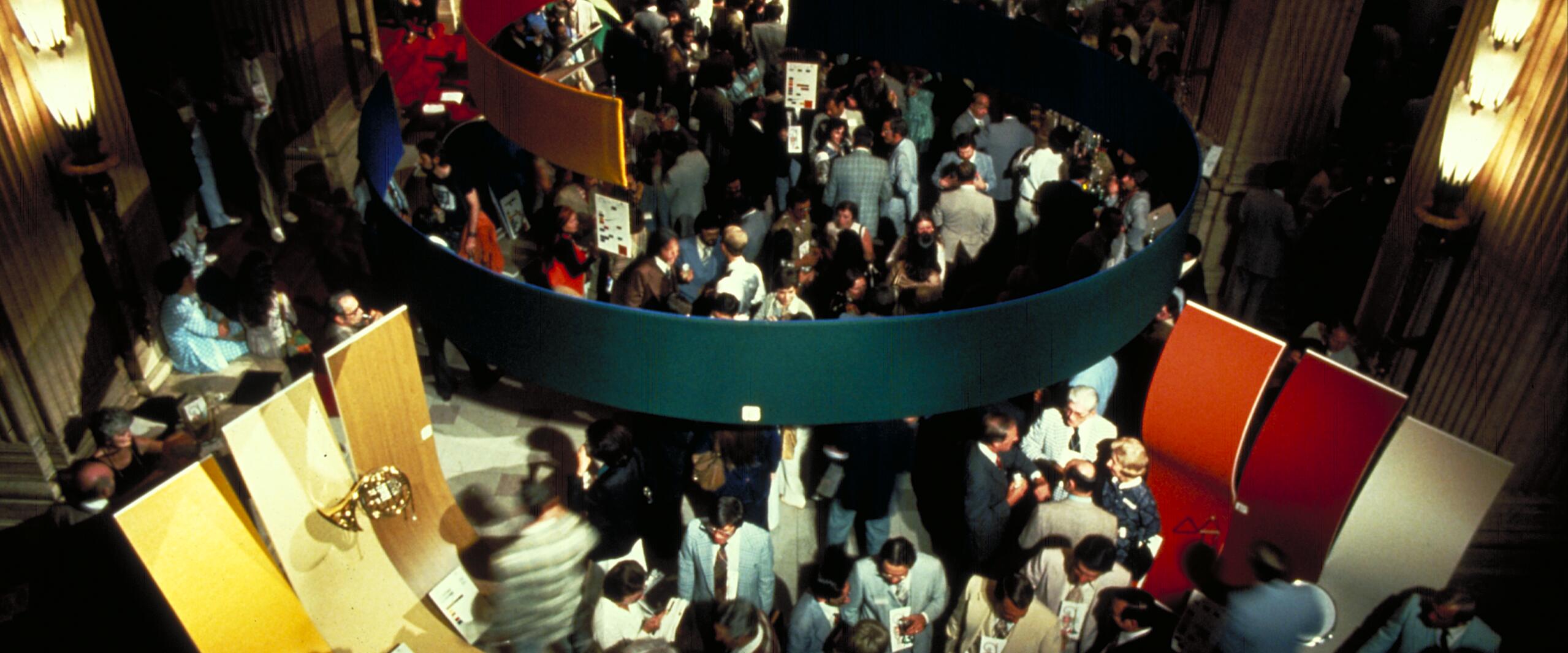 Crowded exhibition hall with people mingling around colorful, curved panels suspended from the ceiling. Warm lighting creates a lively atmosphere.