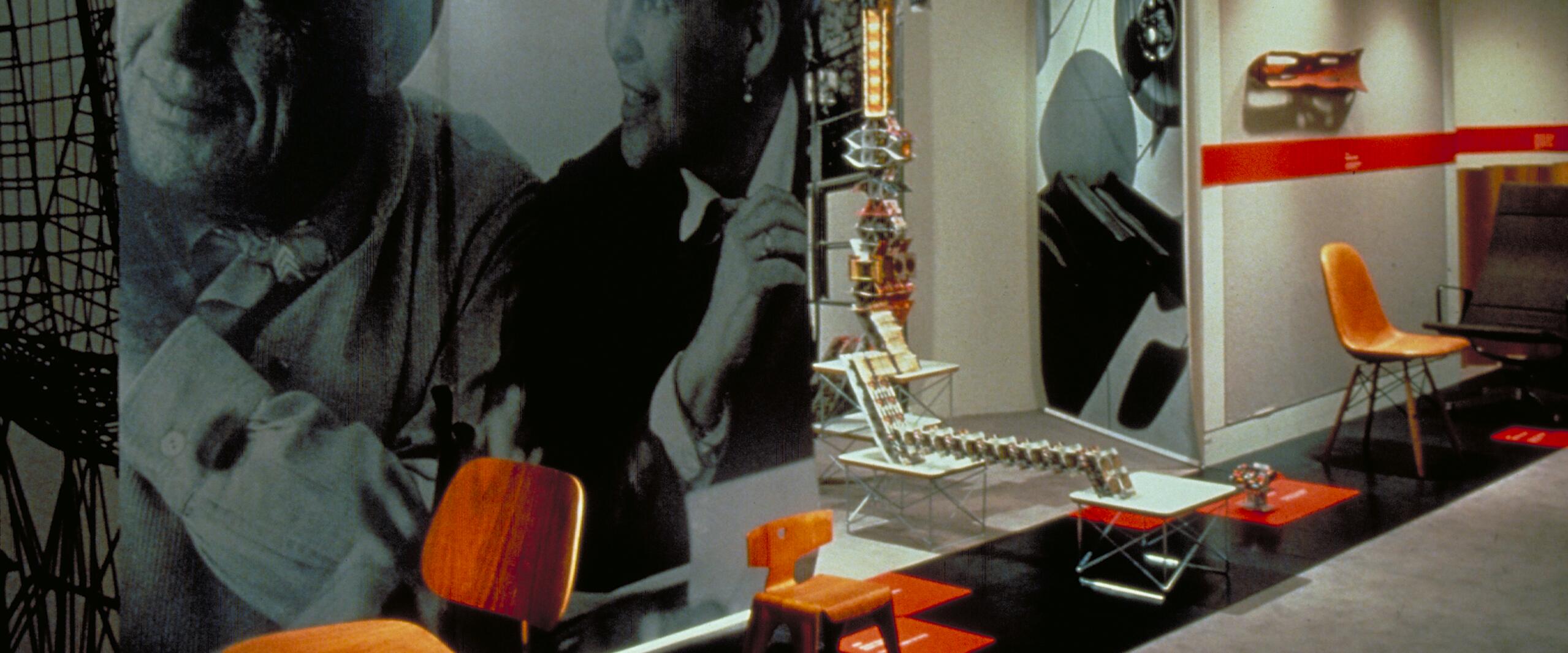 Exhibition space featuring modern chairs and architectural models on display, with two large black-and-white portraits in the background.